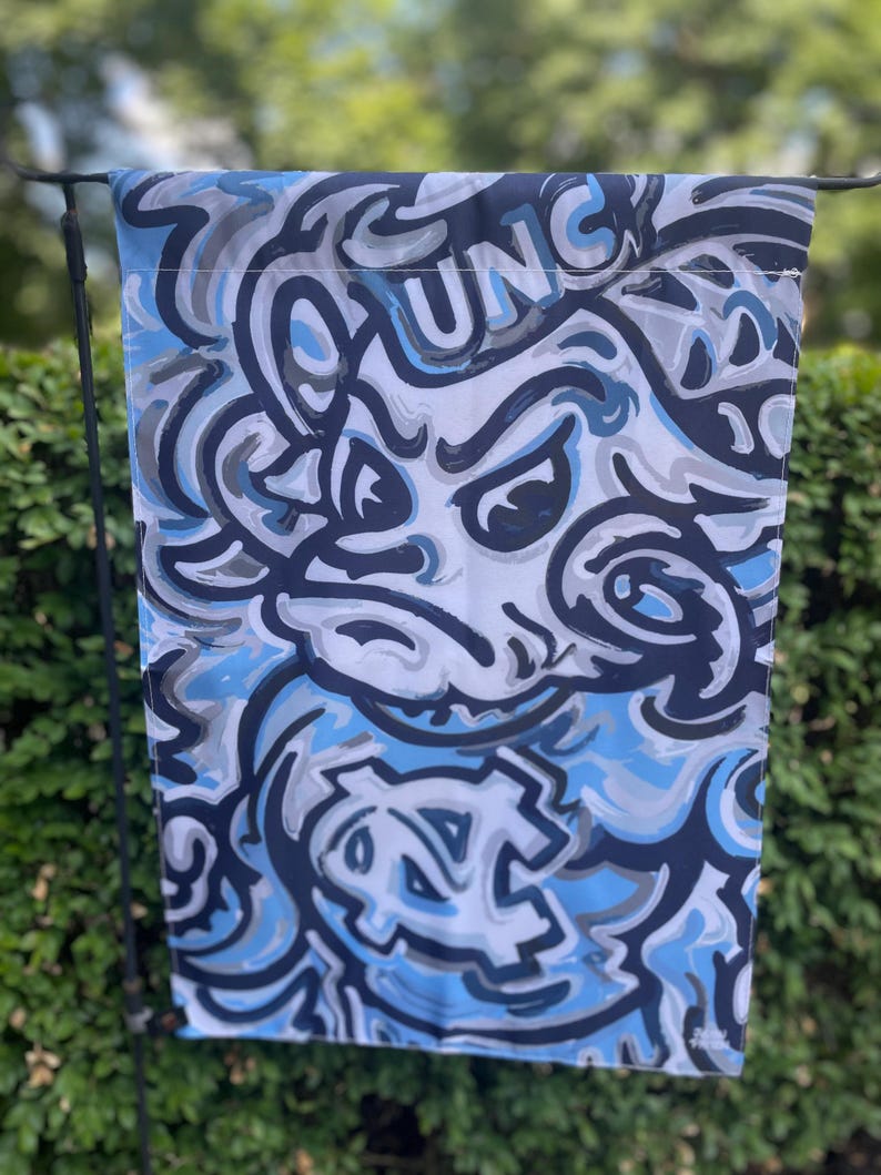 May include: A rectangular flag featuring a stylized design in shades of blue, gray, and white. The design includes the word "UNC" and a graphic of a snarling mascot. The flag is hanging outdoors against a backdrop of green foliage.