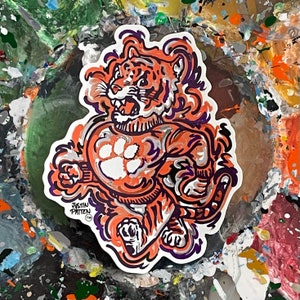 May include: A colorful sticker featuring a cartoon tiger wearing a sweater with a paw print design. The tiger is running and has a fierce expression. The sticker is signed "Justin Patten TM".