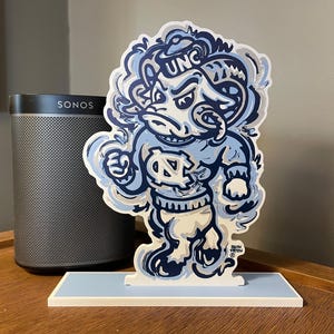 May include: A layered, light blue and white wooden sculpture of a ram mascot, with the letters "UNC" on its head. The sculpture is on a light blue base. A black speaker is in the background.