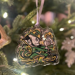 May include: A clear acrylic Christmas ornament featuring the Notre Dame Fighting Irish mascot in green, gold, and black. The ornament is hanging from a pink ribbon against a backdrop of a Christmas tree.