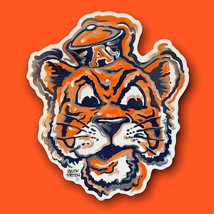 May include: A sticker of an orange and blue cartoon tiger wearing a blue and white hat with the letter "A" on it. The tiger has a mischievous expression and is looking to the right. The sticker is on a bright orange background.
