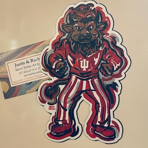 May include: A die-cut sticker featuring a stylized illustration of a bison mascot in a red and white uniform. The bison is in a powerful stance, with the Indiana University logo on its chest. A business card is in the background.