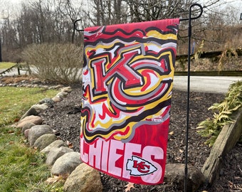 May include: A red, white, and yellow garden flag with the Kansas City Chiefs logo and the word "CHIEFS" in large, white letters. The logo is a stylized "KC" with an arrow pointing to the right. The flag is hanging from a black metal pole.