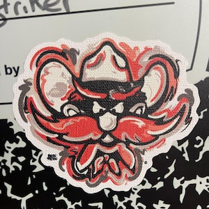 May include: A red, white, and black cartoon mascot with a cowboy hat and a large mustache. The mascot has a fierce expression and is set against a white background.