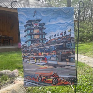 May include: A garden flag featuring an artistic rendering of a race track with a prominent tower and a race car. The flag has a blue sky background with clouds. The race car is orange and red. The artist's signature is in the lower left corner.
