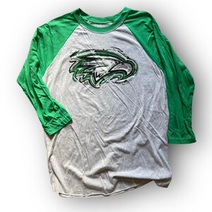 May include: A gray and green baseball tee shirt with a green and black eagle graphic on the front. The shirt has a green three-quarter length sleeve on the left side and a green three-quarter length sleeve on the right side.