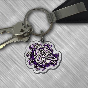 May include: A silver key chain with a purple and white bulldog graphic charm. The charm is attached to a silver ring and a black leather key fob.