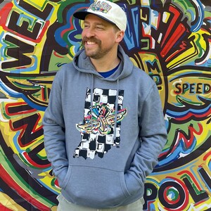 May include: A light blue hoodie with a graphic of the state of Indiana, a checkered flag, and a racing-themed design. The person in the image is wearing a white and blue baseball cap. The background is a colorful mural with the words "Welcome" and "Speedway".