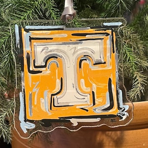 May include: A clear, square Christmas ornament featuring a stylized orange and white letter "T" design with black and light blue accents. The ornament hangs from a silver ribbon against a backdrop of evergreen branches.