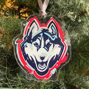 May include: Clear acrylic ornament featuring a Husky dog face in red, white, and blue. The ornament has a white ribbon for hanging and is set against a backdrop of green pine needles. The Husky's face is detailed with a red outline.