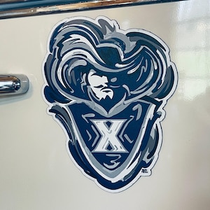 May include: A sticker featuring a stylized design in shades of blue, gray, and white. The design includes a shield shape with a large letter "X" in the center, surrounded by a detailed illustration of a face and flowing hair.