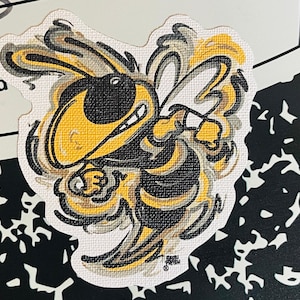 May include: A yellow and black cartoon hornet mascot with a fierce expression. The hornet is drawn in a stylized, almost abstract way, with its wings and body blending together. The mascot is set against a white background.