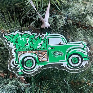 May include: A green and white Christmas ornament shaped like a vintage truck carrying a Christmas tree. The truck has the Philadelphia Eagles logo on the door. The ornament is hanging from a white ribbon.