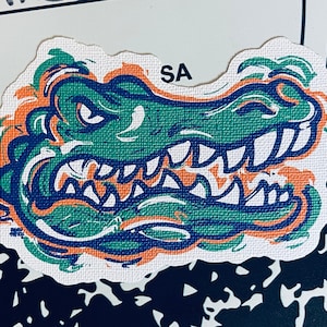 May include: A green and orange alligator head graphic with a white background. The alligator has a toothy grin and is drawn in a stylized, cartoonish style. The text "SA" is visible in the upper right corner of the image.
