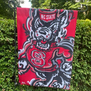 May include: A red garden flag featuring a stylized wolf mascot with the text "NC STATE" at the top. The flag has a bold design with red, black, white, and gray elements, and the number "20" is prominently displayed.