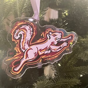 May include: Clear acrylic Christmas ornament featuring a stylized squirrel in shades of pink, maroon, and yellow, with a ribbon for hanging. The squirrel is in a running pose, with a flame-like design around it. The ornament is hanging on a Christmas tree.