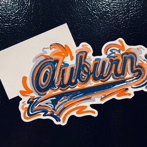 May include: A white sticker with the word "Auburn" written in blue and orange paint splatters. The word is stylized in a bold, brushstroke font.