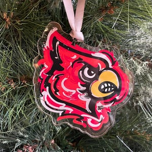 May include: Red, white, and black Christmas ornament featuring a cartoon cardinal bird with a yellow beak. The ornament is made of clear plastic and has a jagged edge.