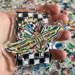 May include: A colorful acrylic keychain featuring a stylized winged design with the text "Indianapolis Motor Speedway". The wings are painted in a rainbow of colors, and the background is a black and white checkered pattern.