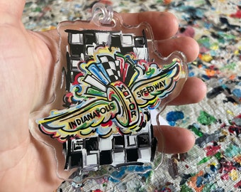 Może przedstawiać: Kolorowy brelok akrylowy z ozdobnym wzorem skrzydeł i tekstem "Indianapolis Motor Speedway". Skrzydła są pomalowane w tęczowych kolorach, a tło stanowi czarno-biały wzór w kratkę.