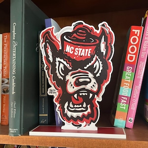 May include: A decorative NC State Wolfpack logo, in red, black, and white, stands on a shelf among books. The logo features the team name and a stylized wolf head. The books include cookbooks and other titles.