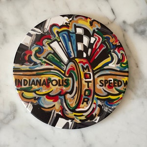 May include: A round, colorful coaster with a stylized image of a wheel and the words "Indianapolis Motor Speedway".