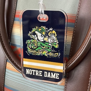May include: A luggage tag with the Notre Dame Fighting Irish logo, featuring a leprechaun in green and gold. The tag is attached to a bag with brown leather handles and a colorful striped pattern.