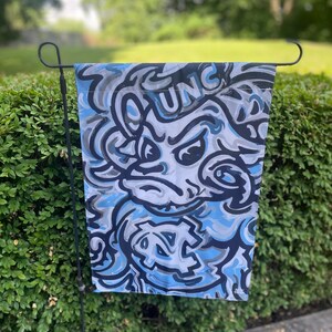 May include: A garden flag featuring a blue, gray, and white abstract design with the letters "UNC" visible. The flag is hanging on a black metal stand in front of a green hedge.