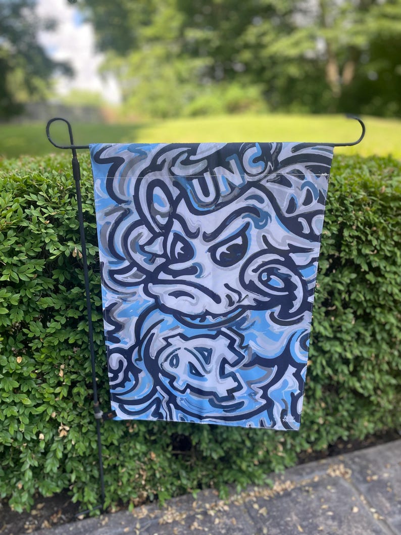 May include: A garden flag featuring a blue, gray, and white abstract design with the letters "UNC" visible. The flag is hanging on a black metal stand in front of a green hedge.