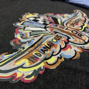 Puede incluir: Un dise&ntilde;o gr&aacute;fico colorido de un ala estilizada con el texto "Indianapolis Motor Speedway" impreso en &eacute;l. El dise&ntilde;o est&aacute; impreso en una camiseta gris oscura.