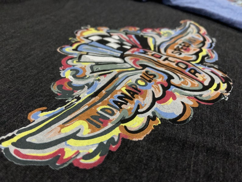 Puede incluir: Un dise&ntilde;o gr&aacute;fico colorido de un ala estilizada con el texto "Indianapolis Motor Speedway" impreso en &eacute;l. El dise&ntilde;o est&aacute; impreso en una camiseta gris oscura.