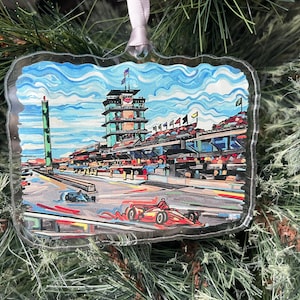 May include: Clear, rectangular Christmas ornament featuring a colorful watercolor painting of a racetrack with race cars. The ornament has a scalloped edge and is suspended by a white ribbon.