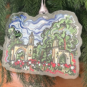 May include: A glass Christmas ornament featuring a stylized illustration of a white building with two arched entrances, surrounded by green trees and red flowers. The sky is blue with white clouds.