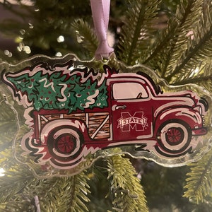 May include: A red truck with a Christmas tree in the bed. The truck has a "STATE M" logo on the side. The truck is a clear acrylic ornament.