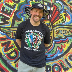 May include: A black t-shirt with the text "Indianapolis Motor Speedway" and a colorful graphic design. The person is wearing a black hat with a checkered pattern and a logo. The background is a colorful mural with the words "Welcome" and "Indianapolis Speedway".