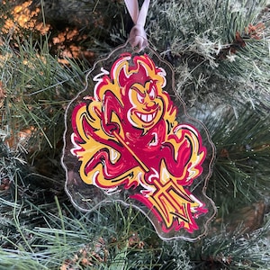 May include: A clear plastic Christmas ornament featuring a red and yellow cartoon devil holding a trident. The devil is surrounded by flames.