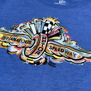 Puede incluir: Una camiseta azul con un dise&ntilde;o gr&aacute;fico colorido del logotipo del Indianapolis Motor Speedway. El logotipo presenta un ala estilizada con las palabras "Indianapolis Motor Speedway" y el nombre del artista "Justin Patten".