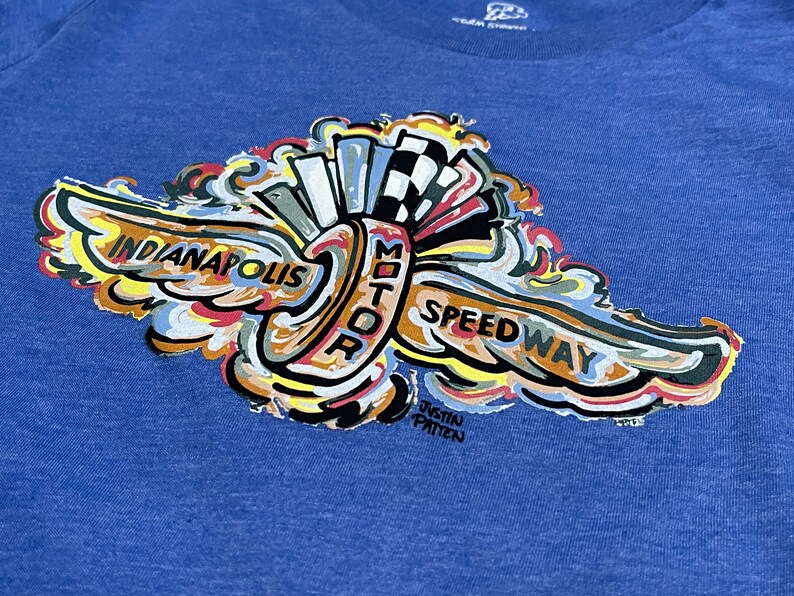Puede incluir: Una camiseta azul con un dise&ntilde;o gr&aacute;fico colorido del logotipo del Indianapolis Motor Speedway. El logotipo presenta un ala estilizada con las palabras "Indianapolis Motor Speedway" y el nombre del artista "Justin Patten".