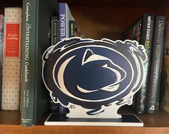 May include: A Penn State Nittany Lions logo bookend in navy blue and white, standing on a shelf with cookbooks and other books. The bookend has the artist's signature, Justin Patten.
