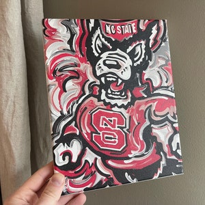 May include: A canvas painting featuring the NC State Wolfpack logo. The artwork showcases a snarling wolf in red, black, and white brushstrokes. The words "NC STATE" are at the top, and the letter "S" is prominent.