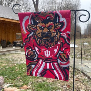 May include: A red and white garden flag featuring a cartoon bull mascot wearing a red jersey with the letter "I" and the Indiana University logo. The flag is hanging on a black metal stand.