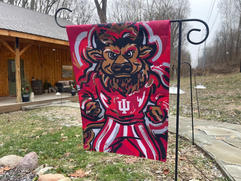 May include: A red and white garden flag featuring a cartoon bull mascot wearing a red jersey with the letter "I" and the Indiana University logo. The flag is hanging on a black metal stand.