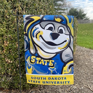 May include: A colorful garden flag featuring a cartoon dog mascot with a blue and yellow paint splatter design. The flag reads "STATE" and "SOUTH DAKOTA STATE UNIVERSITY".