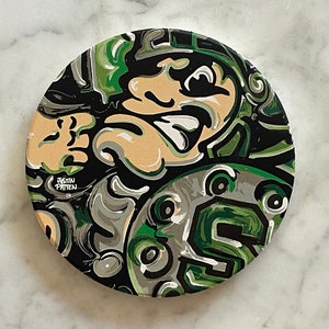 May include: A round coaster with a black, green, and white abstract design. The design features a face with a large nose and mouth. The text "Justin Patten" is visible in the lower left corner.