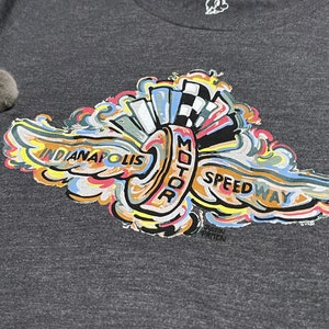 Puede incluir: Una camiseta gris con un dise&ntilde;o gr&aacute;fico colorido de un neum&aacute;tico estilizado con alas y el texto "Indianapolis Motor Speedway".