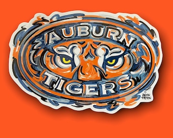 May include: An oval-shaped sticker with the words "AUBURN TIGERS" in white, orange, and navy blue. The design features a tiger's face with yellow eyes. The sticker has a white border and is set against an orange background.