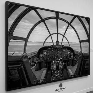 May include: Black and white photograph of an aircraft cockpit. The view from the cockpit shows a runway and a cloudy sky. The photo is framed in a black frame.