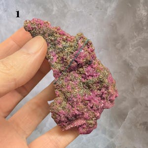 May include: A close-up of a mineral specimen with a vibrant pink hue, speckled with patches of gold and dark green. The rough, textured surface of the stone is held in a hand, with the number "1" visible in the upper left corner.