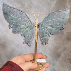 May include: A decorative butterfly sculpture with a grey and green stone-like appearance. The butterfly has a gold-coloured metal body and antennae, and is mounted on a gold-coloured metal stand. The wings have a textured surface.