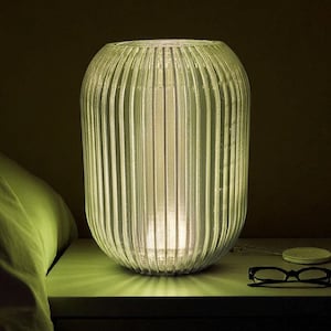 Könnte beinhalten: Eine gerippte, zylindrische Lampe strahlt ein sanftes Licht auf einem Nachttisch aus. Daneben befindet sich ein Bett mit einem weißen Kissen. Eine Brille, ein kabelloses Ladegerät und ein Buch liegen ebenfalls auf dem Tisch.